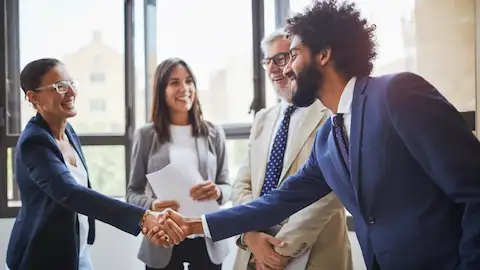 Quatro profissionais em um ambiente corporativo iluminado, sorrindo enquanto dois deles apertam as mãos em um gesto de cumprimento.